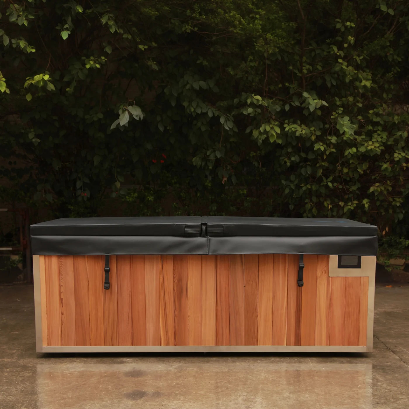 A rectangular outdoor hot tub or spa with a wood-look brown front panel, light beige trim, and a dark gray cover, photographed on a patio with dense green foliage in the background.
